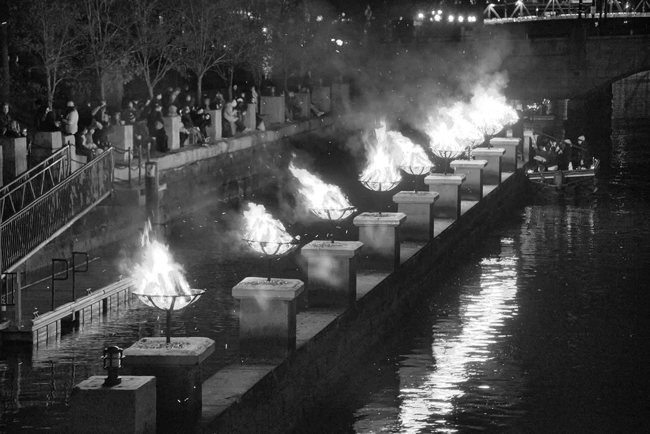 A black and white photo of the braziers along the Providence River being lit as part of the WaterFire festival. Cianci was a key figure in the creation of the festival and was instrumental in the uncovering of the Providence River and the establishment of Providence as a city dedicated to arts and culture.